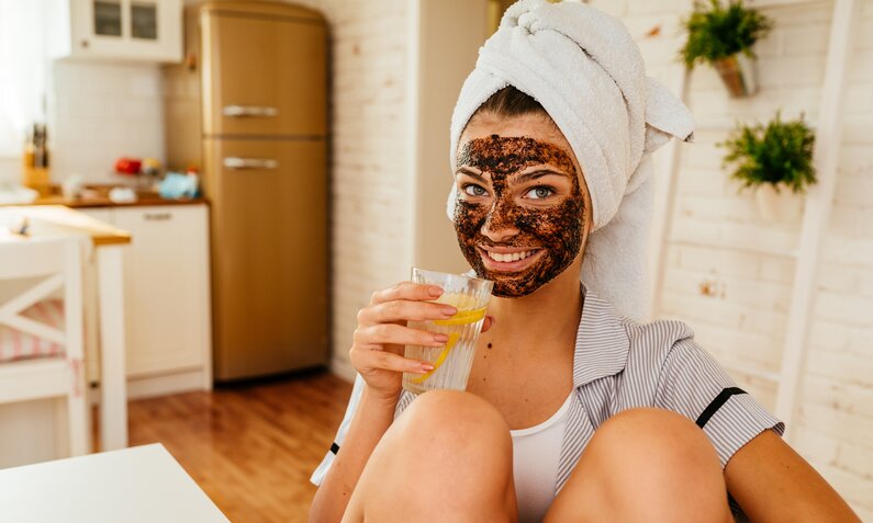 Junge Frau mit Kaffeemaske lächelt, hält Saftglas in heller Küche. | © Getty Images/ MelkiNimages