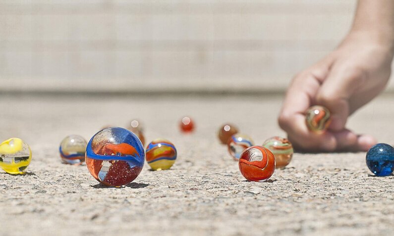 Glänzende Murmeln in lebhaften Farben liegen verstreut auf hellem Beton, eine Hand greift eine. | © Getty Images / Moment / Alicia Llop