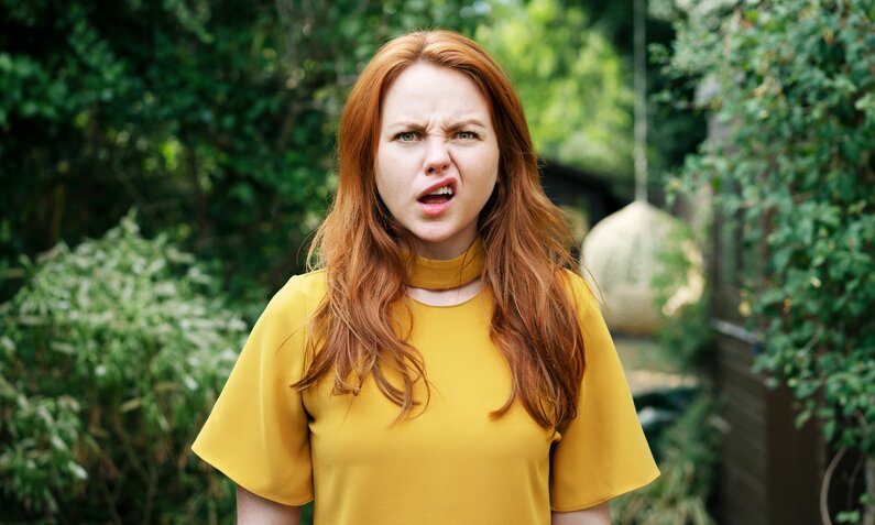 Junge Frau mit roten Haaren zeigt überraschten Gesichtsausdruck in einem gelben Oberteil vor grünem Gartenhintergrund. | © Getty Images/ Tim Robberts