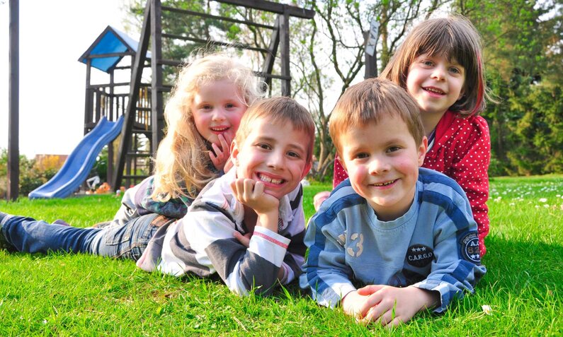 Gruppe fröhlicher Kinder spielt auf einer Wiese, Spielgeräte und Bäume im Hintergrund. | © Kathleen Rekowski/stock.adobe.com 