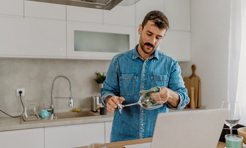 Mann poliert Glas in Küche, steht dabei vor einem Küchentresen und trägt ein blaues Jeanshemd. | © Getty Images / milan2099