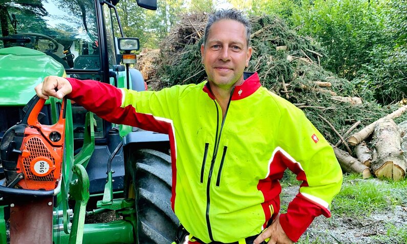 Ein lächelnder Mann in neongelber Arbeitsjacke und roter Hose lehnt an einem grünen Traktor, Motorsäge in der Hand. | © RTL