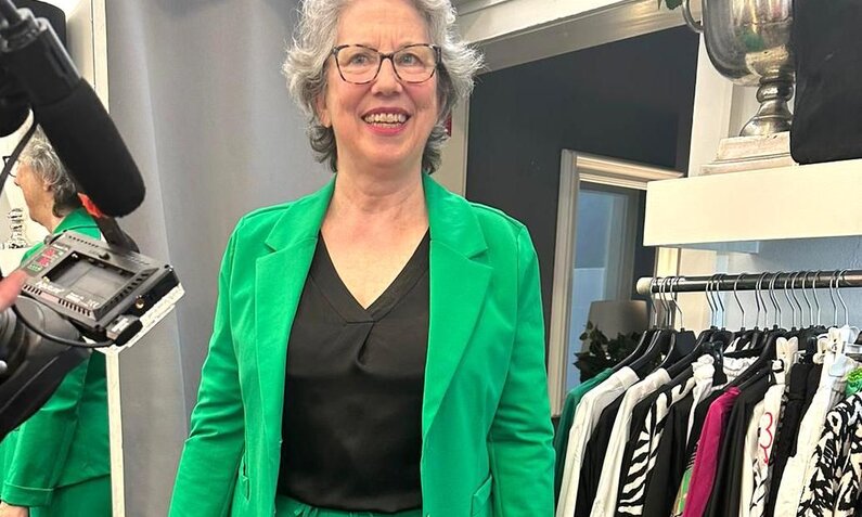 Ältere Dame mit grauem Haar lächelt in grünem Blazer vor Kleiderständern und Spiegel. | © RTL/Constantin Ent.