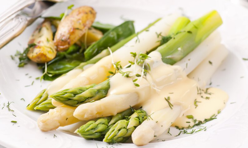 Spargel in Nahaufnahme begossen mit Sauce Hollandaise, angerichtet auf einem weißen Teller, daneben Kartoffeln. | © Getty Images / hlphoto