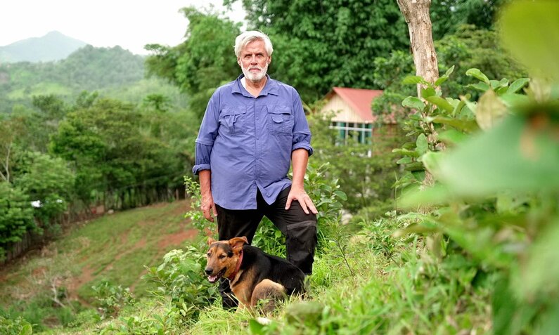 Älterer Mann mit grauem Bart in blauem Hemd steht neben braunem Hund in grüner Berglandschaft. | © RTL/Maria Heymer