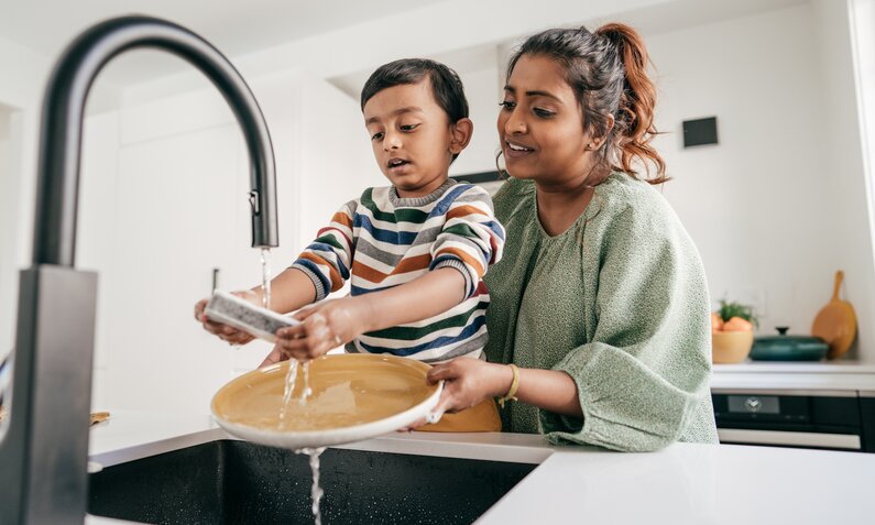 Eine junge Frau hält einen kleinen Jungen an ein Waschbecken und der Junge hält einen Teller unter laufendes Wasser. | © Getty Images / kate_sept2004