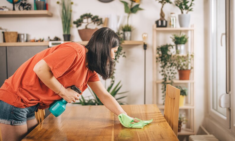 Frau mit braunen Haaren wischt mit einem Tuch über einen Holztisch, dahinter viele Pflanzen. | © Getty Images / freemixer