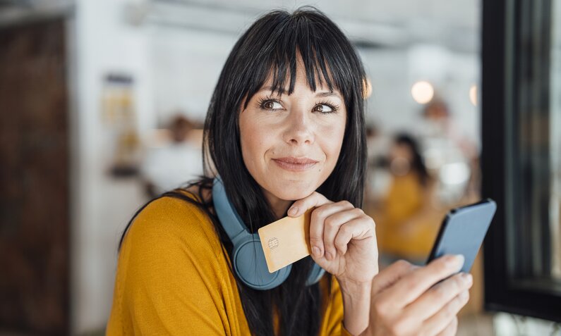 Eine junge Frau mit langen schwarzen Haaren und gelbem Pullover hält eine Kreditkarte und ein Smartphone, lächelnd in einem Café. | © Getty Images/Westend61