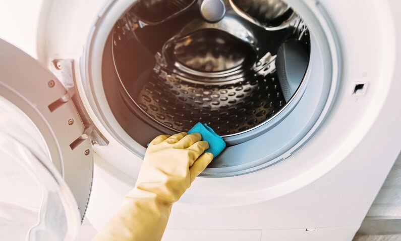 Hände in Putzhandschuhen wischen mit einem Tuch über das Innere einer Waschmaschine. | © Getty Images / 	Cris Cantón