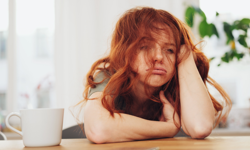 Eine junge Frau mit langen roten Haaren stützt ihren Kopf auf den Armen auf einen Tisch und wirkt erschöpft. | © Adobe Stock/ contrastwerkstatt