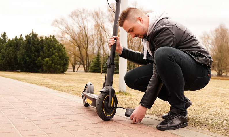 Junger Mann von der Seite in dunkler Lederjacke und Jeans kniet vor schwarzem E-Scooter und hält ihn mit einer Hand fest, mit der anderen Hand hält er eine kleine schwarze Akku-Luftpumpe an das Vorderrad, auf hellem Steinweg vor Grünfläche | © Adobe/ brillianata