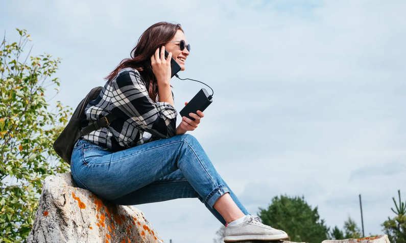 Junge Frau von der Seite lachend auf hellen Felsen vor blauem Himmel sitzend in Jeans und Flanellhemd hält dunkles Smartphone ans Ohr und in der anderen Hand eine schwarze Powerbank | © Adobe/ LesdaMore