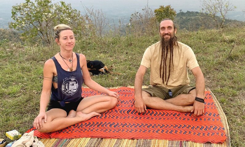 Eine blonde Frau und ein Mann mit langen Dreadlocks sitzen meditierend auf einer orangefarbenen Decke in einer grünen Hügellandschaft. | © RTL