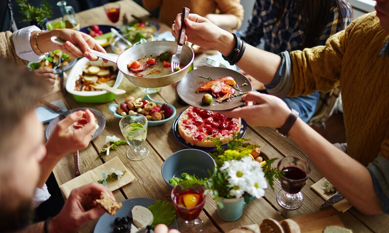 Eine Gruppe von Freunden genießt ein gemeinsames Essen an einem Holztisch mit verschiedenen Speisen und Getränken. | © Adobe Stock/ pressmaster