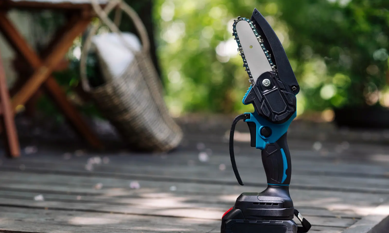 Blau schwarze Mini-Kettensäge aufgestellt auf Holzveranda, im Hintergrund verschwommen Stuhl- und Tischbeine und rechts grüne Bäume und Pflanzen | © Adobe/ YarikL