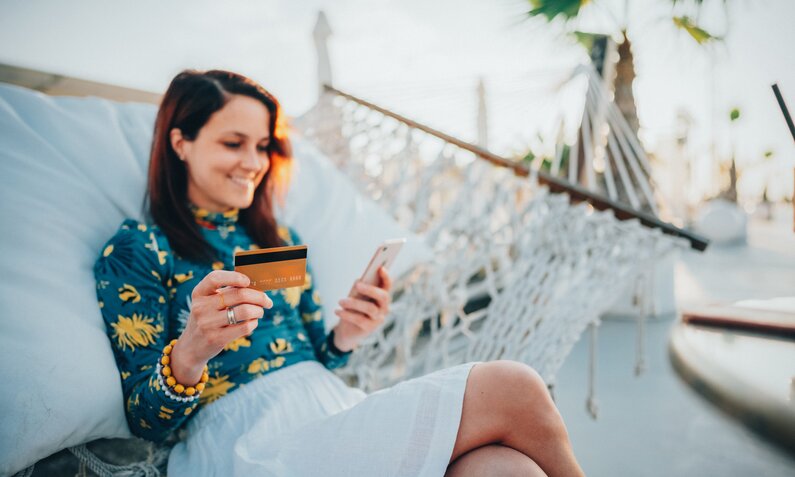 Eine junge Frau mit braunen Haaren sitzt lächelnd in einer Hängematte, hält eine Kreditkarte und ein Smartphone. | © Getty Images/martin-dm