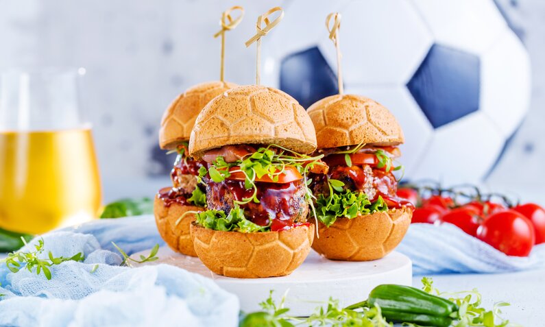 Drei Burger reicht belegt mit Fleisch. Soße, Salat und Käse, Burgerbuns in Form eines Fußballes und dahinter ein Fuißball. | © Adobe Stock / karepa