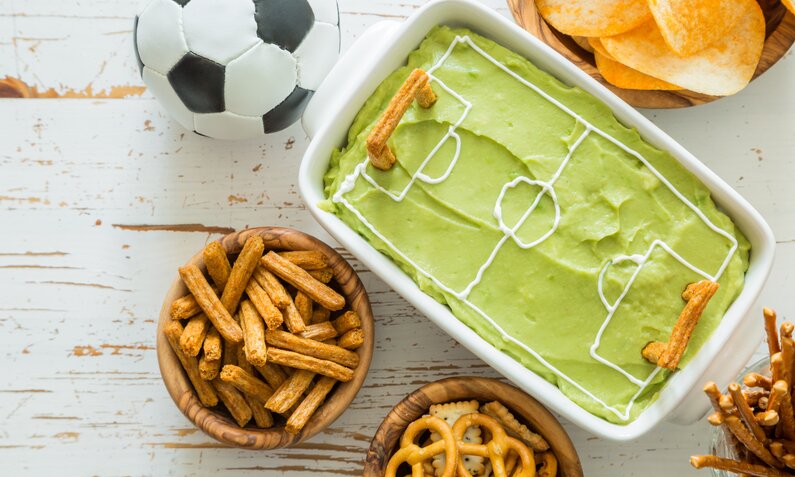 Auflaufform gefüllt mit Guacamole im Look eines Fußballfeldes, daneben Chips, Fußball, Snacks. | © Adobe Stock / anaumenko