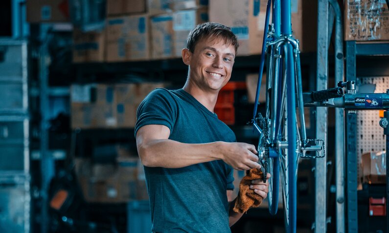 Dennis Mojen steht lächelnd  in einer Werkstatt und repariert eine Fahrrad in einer Szene aus dem Netflix-Film "Spieleabend" | © Netfllix/Sasha Ostrov