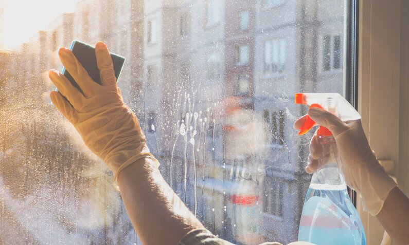 Hände in gelben Gummihandschuhen putzen mit Schwamm und Reinigungsmittel ein Fenster in der Morgensonne. | © Adobe Stock/ sablinstanislav