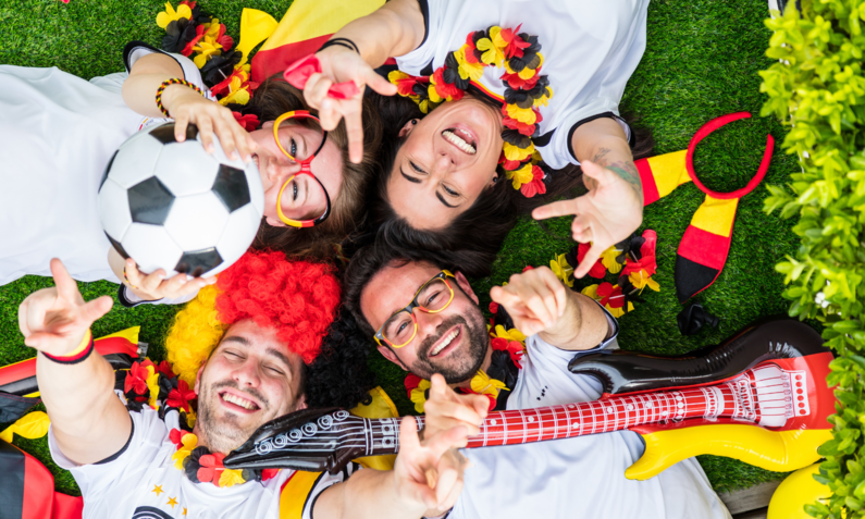 Vier jubelnde Fußballfans in Deutschlandtrikots liegen lachend auf dem Rasen, geschmückt mit Girlanden in Schwarz-Rot-Gold. | © Adobe Stock/ karepa