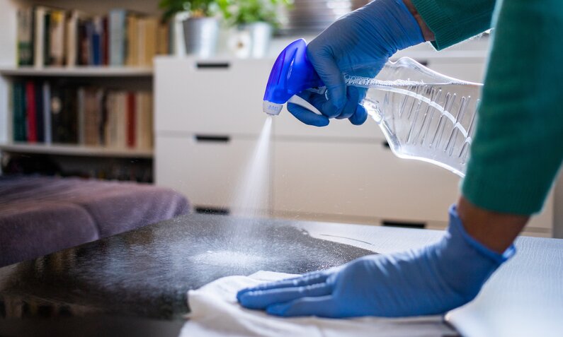 Eine Person mit blauen Handschuhen sprüht Reinigungsmittel auf einen Tisch in einem gemütlichen Wohnzimmer. | © Adobe Stock/ Melinda Nagy