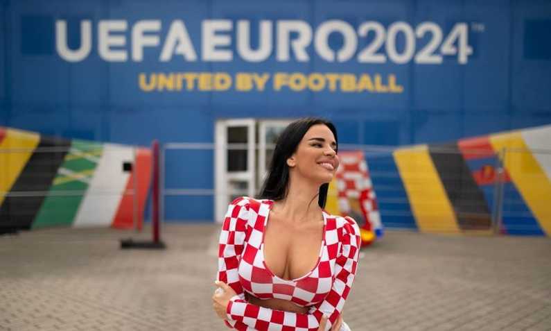 Junge Frau mit schwarzen Haaren und rot-weiß kariertem Oberteil lächelt vor UEFA EURO 2024-Banner. | © knolldoll/Instagram/Screenshot