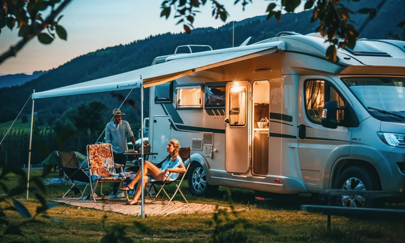 Weißes Wohnmobil schräg von der Seite in Abenddämmerung in Natur mit ausgefahrener Markise und einem jungen Paar, das auf Campingmöblen sitzt | © Fxquadro/ Adobe Stock