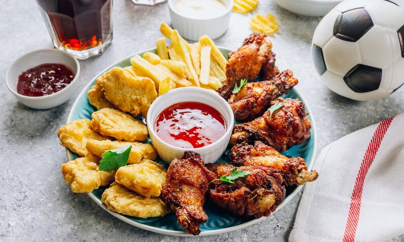 Gemischte Snackplatte mit Chicken Wings, Chicken Nuggets und Pommes, daneben ein Fußball, Dips und Getränke. | © Getty Images / Irina Kozmova