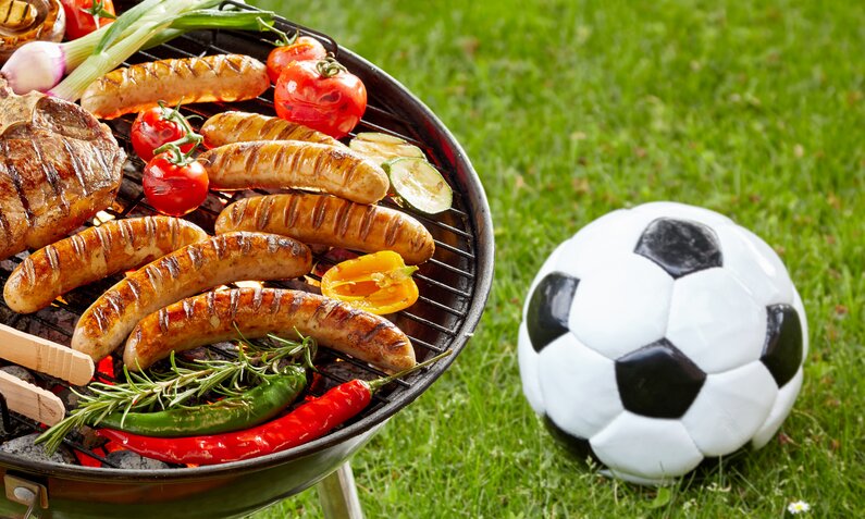 Auf einem Grill brutzeln Bratwürste, Gemüse und Steaks, daneben liegt ein Fußball auf grünem Rasen. | © Adobe Stock/ exclusive-design