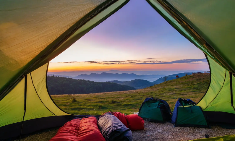 Ausblick aus geöffnetem grünem Zelt in dem ein paar zusammengerollte Schlafsäcke und eine Tasche liegen auf Berglandschaft mit Sonnenaufgang | © Florence/ Adobe Stock