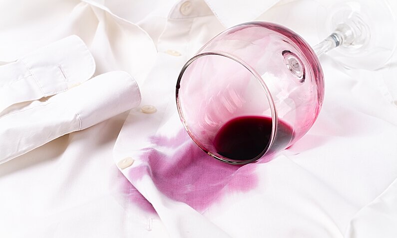 Ein umgekipptes Weinglas mit rotem Wein hat einen großen Fleck auf einem weißen Hemd hinterlassen. | © Adobe Stock/ Gala_Didebashvili