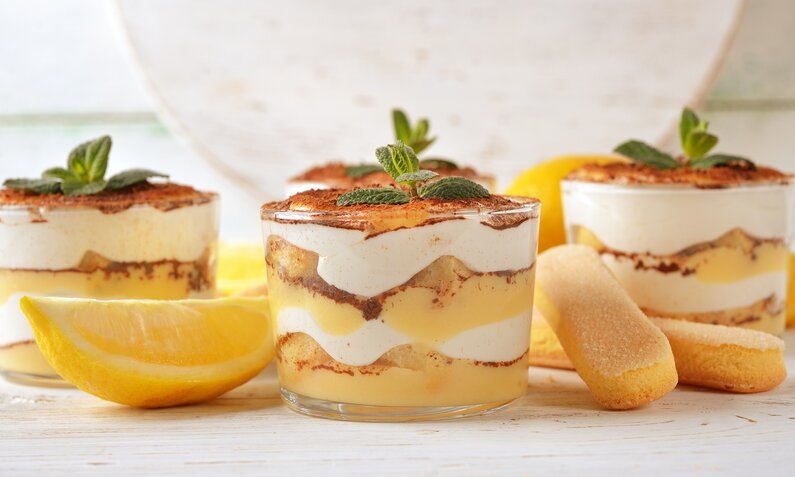 Zitronentiramisu angerichtet in drei Glas-Dessertgläsern, daneben frische Zitronenscheiben und Löffelbiskuits. | © Getty Images / olvas