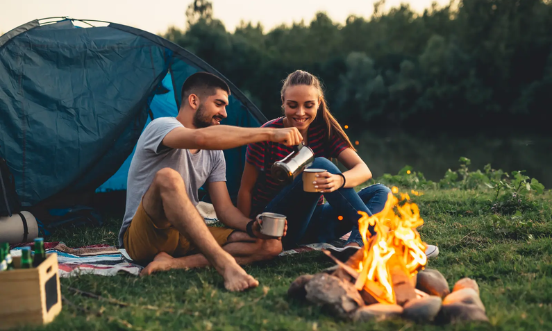 Junges Pärchen sitzt lächelnd auf Wiese hinter Lagerfeuer vor Zelt; Mann hält Becher in der einen und Kanne in der anderen Hand und gießt Frau mit Becher Kaffee ein | © cherryandbees/ Adobe Stock