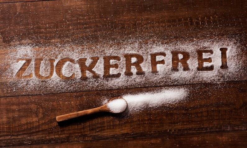 Das Wort "zuckerfrei" ist in Zucker auf einem dunklen Holztisch geschrieben, daneben ein Holzlöffel mit Zucker. | © Adobe Stock/ nito