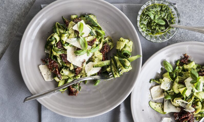 Zucchini-Pasta mit getrockneten Tomaten, Parmesan, angerichtet auf blauem Teller. | © Getty Images / 	HealthyLauraCom