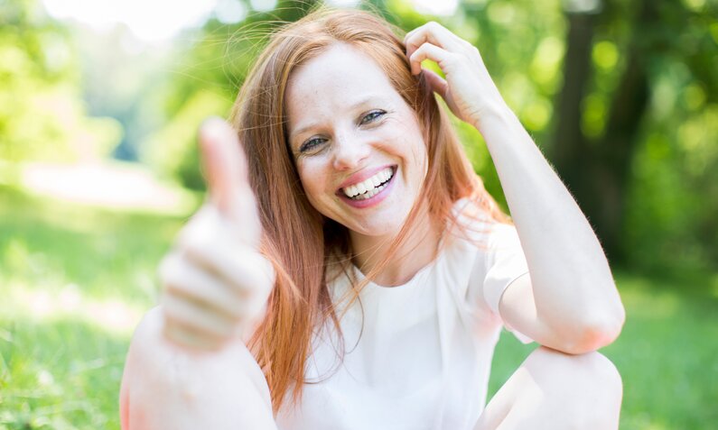 Lachende Frau mit roten Haaren im weißen Shirt gibt im sonnigen Park ein Daumen-hoch-Zeichen. | © Adobe Stock/ drubig-photo