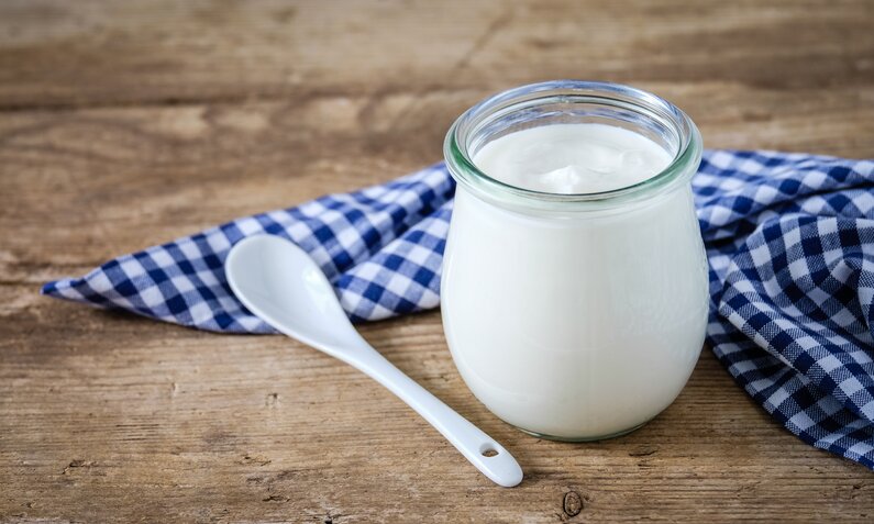 Ein Glas Joghurt steht neben einem weißen Löffel und einem blau-weiß karierten Tuch auf einer rustikalen Holzfläche. | © Adobe Stock/ ji_images