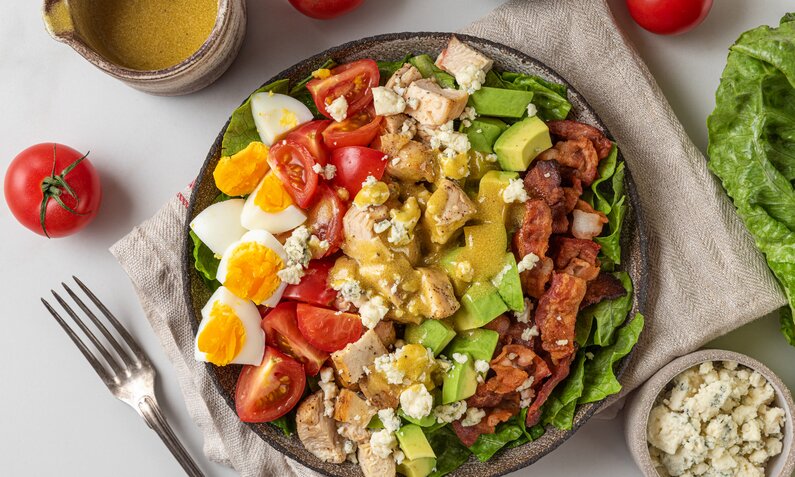 Angerichteter Salat mit Ei, Bacon, Pute, Tomaten, Fetakäse, Romanasalat in einer Schüssel mit Senfdressing. | © Getty Images / samael334