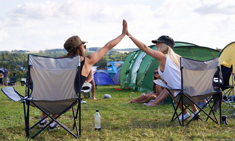 Zwei junge Frauen geben sich lachend High-Five auf Campingstühlen, umgeben von Zelten und Wiese unter einem bewölkten Himmel. | © Adobe Stock/ Julie Francoeur/peopleimages.com