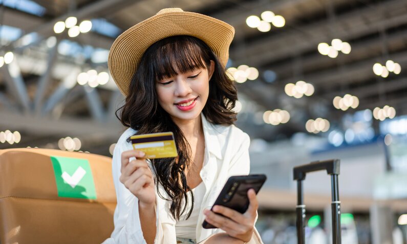 Junge Frau mit Strohhut und langen dunklen Haaren ist am Flughafen und hält lächelnd eine Kreditkarte und ein Smartphone in der Hand. | © Getty Images/Twenty47studio