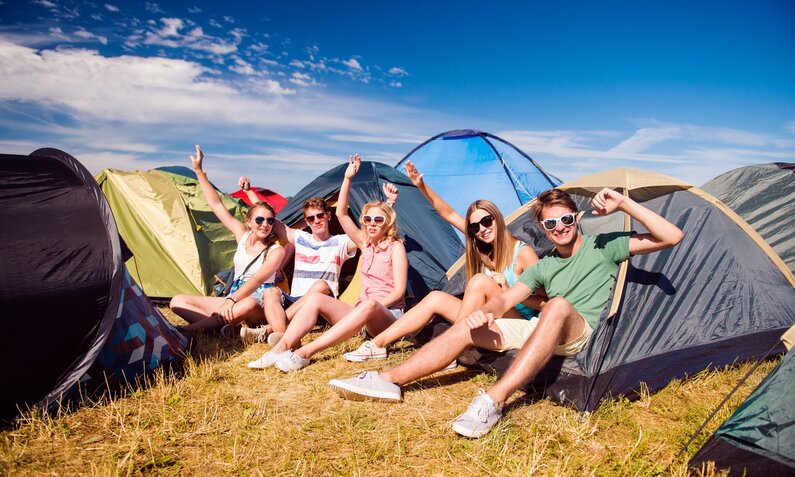 Fünf junge Menschen sitzen lachend vor Zelten auf einer Wiese und winken in die Kamera, unter blauem Himmel. | © Adobe Stock/ Halfpoint