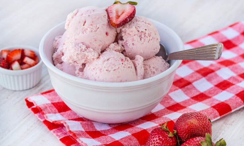 Drei Erdbeereis-Kugeln in einer weißen Schüssel angerichtet, daneben frische Minze und frische Erdbeeren. | © Getty Images / 	carolmellema