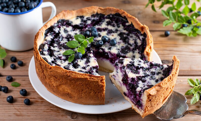 Angeschnittener Blaubeerkuchen, daneben frische Blaubeeren und Kräuter. | © Getty Images / Qwart