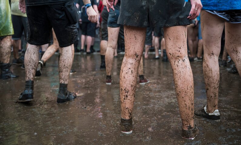 Verschmutzte Beine von Festivalbesuchern in Shorts und Sneakers stehen auf schlammigem Boden. | © Adobe Stock/ Dziurek