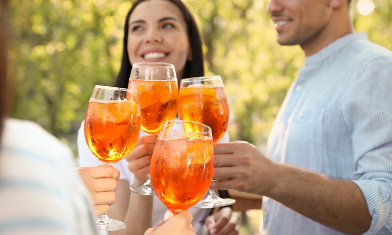 Eine Gruppe fröhlicher Freunde stößt mit Aperol Spritz in einem sonnigen Park an. | © Adobe Stock/ New Africa