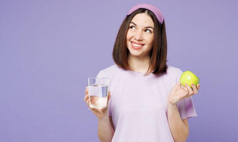 Eine junge Frau mit braunen Haaren und lila Stirnband hält lächelnd ein Glas Wasser und einen grünen Apfel vor lila Hintergrund. | © Adobe Stock/ ViDi Studio