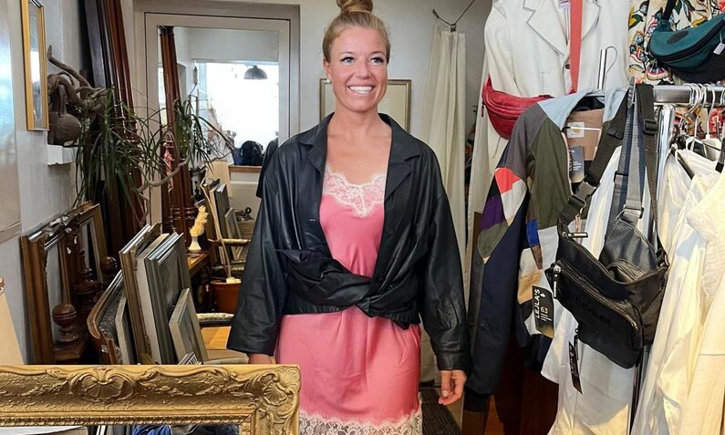 Blonde Frau mit hochgestecktem Haar trägt ein rosa Spitzenkleid und schwarze Lederjacke, umgeben von Vintage-Möbeln und Kleidern. | © RTL/Constantin Ent.