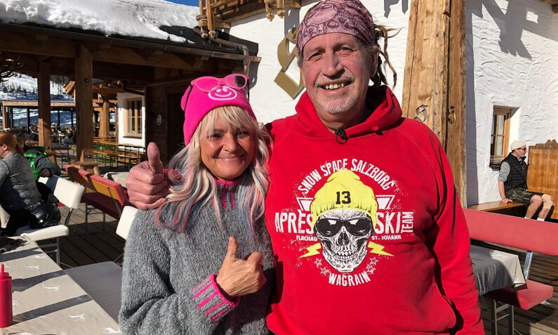 Ein Mann in rotem Hoodie und eine Frau mit rosa Mütze stehen bei Sonnenschein vor einer rustikalen Skihütte. | © RTL/99pro media