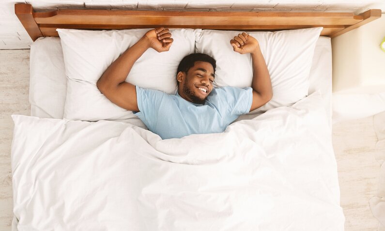 Glücklicher Mann mit kurzem Bart liegt in einem weißen Bett, streckt die Arme und lächelt entspannt. | © Adobe Stock/ Prostock-studio